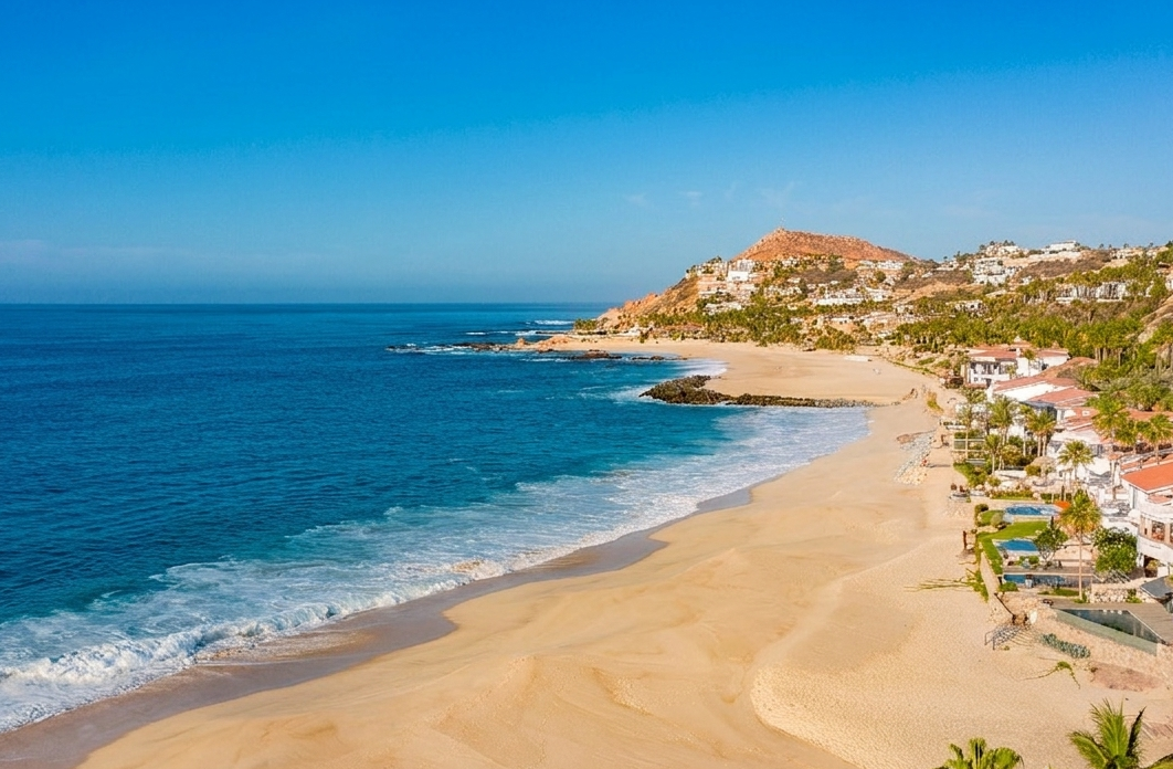 A vibrant, high-angle aerial view of a sweeping crescent beach with golden sand meeting a deep blue ocean. Bright white surf rolls onto the shoreline, while upscale coastal villas with red-tiled roofs, swimming pools, manicured lawns, and palm trees line
